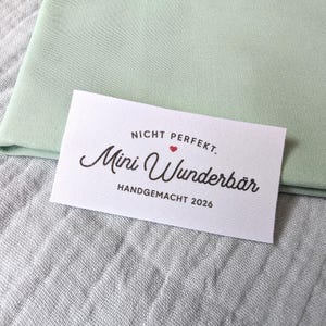 Könnte beinhalten: Ein rechteckiges weißes Stoffetikett mit schwarzem Text, der NICHT PERFEKT. Mini Wunderbär HANDGEMACHT 2026 lautet, sowie einem kleinen roten Herzsymbol, platziert auf einem gefalteten hellen salbeigrünen Stoff.