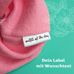Könnte beinhalten: Nahaufnahme eines rosa Stoffes mit einem angenähten weißen Etikett. Das Etikett trägt den Text "outfit of the day" in schwarzer Schrift.
