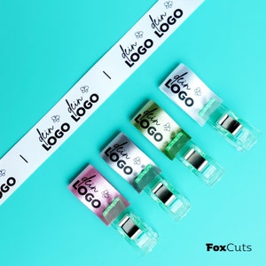 Könnte beinhalten: Vier bunte Stofflabels mit dem aufgedruckten Text "dein LOGO". Die Etiketten sind an durchsichtigen Plastikclips befestigt.