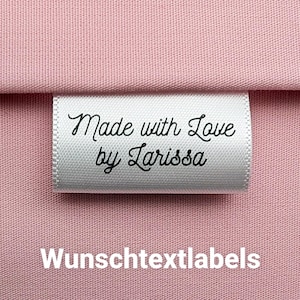 Könnte beinhalten: Weißes Satin-Etikett mit schwarzem Text, der "Made with Love by Larissa" lautet. Das Etikett befindet sich auf einem rosa Stoffhintergrund. Der Text "Wunschtextlabels" ist unter dem Etikett gedruckt.