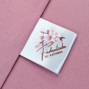 Könnte beinhalten: Weißes Satin-Etikett mit einem floralen Design und dem Text "Fadenliebe by Kathrin" in goldener Schrift.