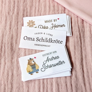 Könnte beinhalten: Drei weiße Stoffetiketten mit Text und Illustrationen. Auf dem oberen Etikett steht "MADE BY Dika Heimer". Auf dem mittleren Etikett steht "FADEN & LIEBE Oma Schildkröte HANDGEMACHT". Auf dem unteren Etikett steht "GESTRICKT VON Andrea Schönwetter". Die Etiketten liegen auf einem rosa Stoff.
