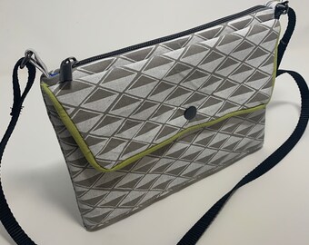 May include: A crossbody bag featuring a gray and white geometric triangle pattern, a lime green fabric trim along the front flap, a black snap button closure, a black zipper top, and a thin black adjustable shoulder strap made of woven nylon.