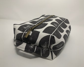 May include: A rectangular fabric toiletry bag featuring a black and white geometric pattern of semi-circles. The bag includes a brass-toned metal zipper closure running along the top and a small fabric handle attached to one end of the pouch.