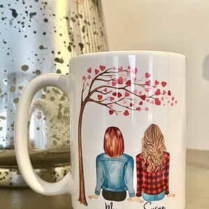 May include: White ceramic mug with a design of two women sitting under a tree with pink hearts. The text "Mom" and "Susan" is printed below the women.
