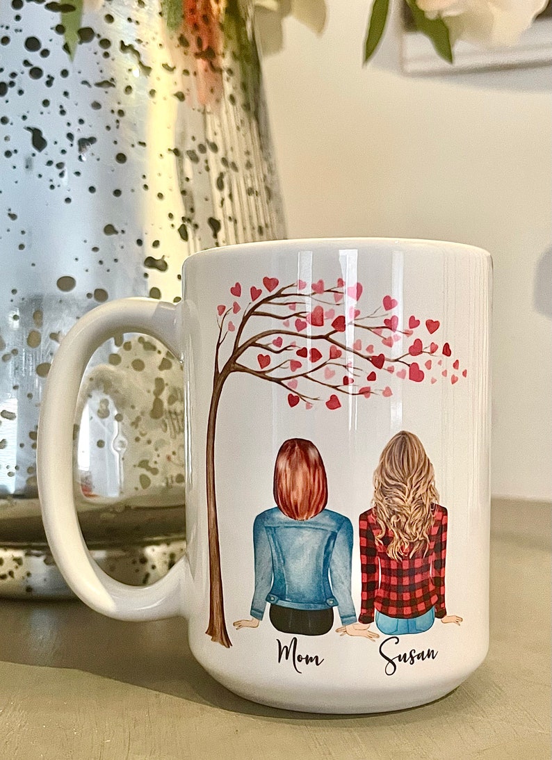 May include: White ceramic mug with a design of two women sitting under a tree with pink hearts. The text "Mom" and "Susan" is printed below the women.