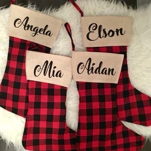May include: Four Christmas stockings featuring a red and black buffalo plaid pattern. Each stocking has a tan cuff with a name printed in black script: Angela, Elson, Mia, and Aidan. Each stocking includes a red and black plaid hanging loop.
