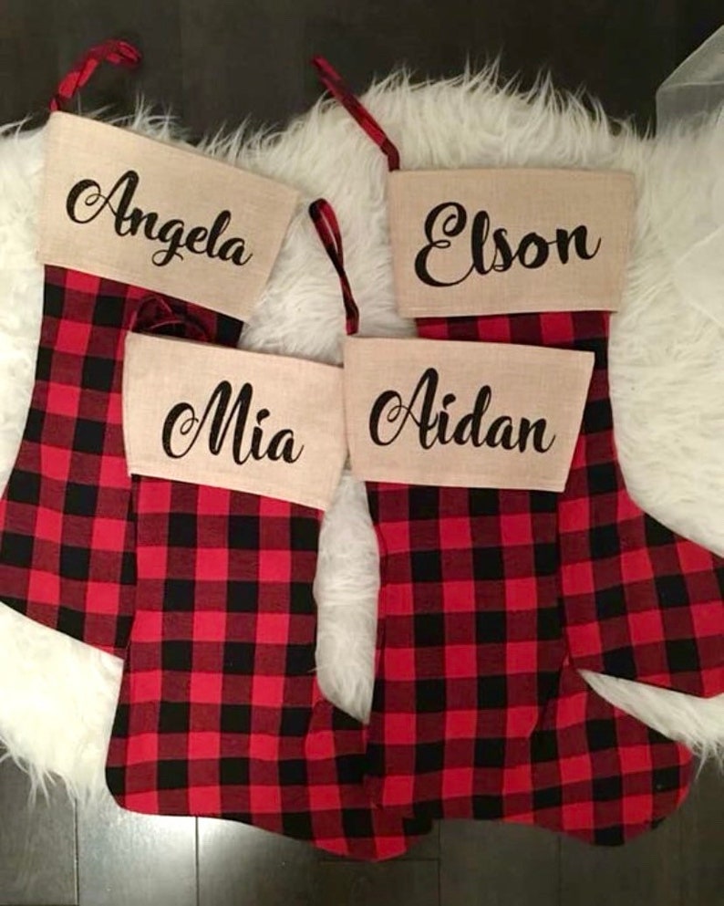 May include: Four Christmas stockings featuring a red and black buffalo plaid pattern. Each stocking has a tan cuff with a name printed in black script: Angela, Elson, Mia, and Aidan. Each stocking includes a red and black plaid hanging loop.