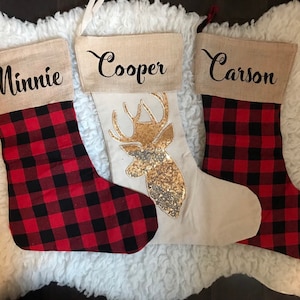 May include: Three Christmas stockings with burlap cuffs. Two stockings feature a red and black buffalo plaid pattern with the names Minnie and Carson printed in black. The center stocking is cream with a gold sequin deer head design.
