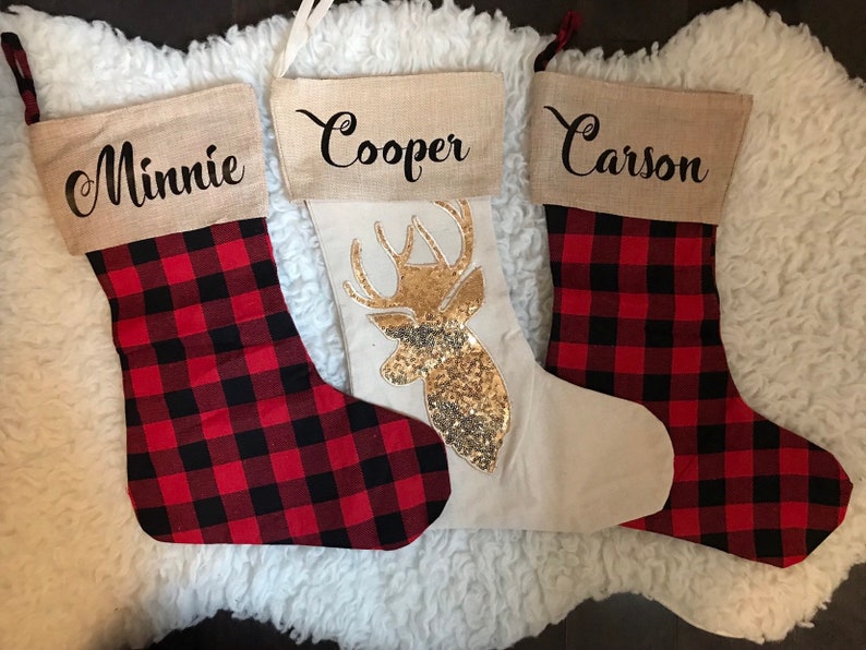 May include: Three Christmas stockings with burlap cuffs. Two stockings feature a red and black buffalo plaid pattern with the names Minnie and Carson printed in black. The center stocking is cream with a gold sequin deer head design.