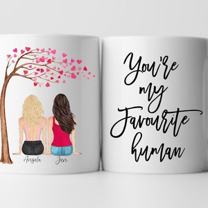 May include: White ceramic mug with a black design that says "You're my favourite human". The other side of the mug features a cartoon illustration of two women sitting under a tree with pink hearts. The women are labeled "Angela" and "Jen".