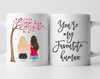 May include: White ceramic mug with a black design that says "You're my favourite human". The other side of the mug features a cartoon illustration of two women sitting under a tree with pink hearts. The women are labeled "Angela" and "Jen".