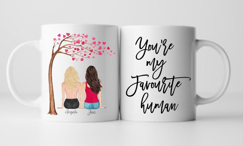 May include: White ceramic mug with a black design that says "You're my favourite human". The other side of the mug features a cartoon illustration of two women sitting under a tree with pink hearts. The women are labeled "Angela" and "Jen".