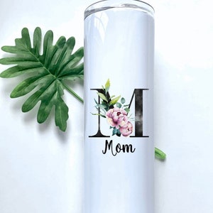 May include: White stainless steel tumbler with a black letter "M" decorated with pink and green floral accents. The word "Mom" is printed below the letter.