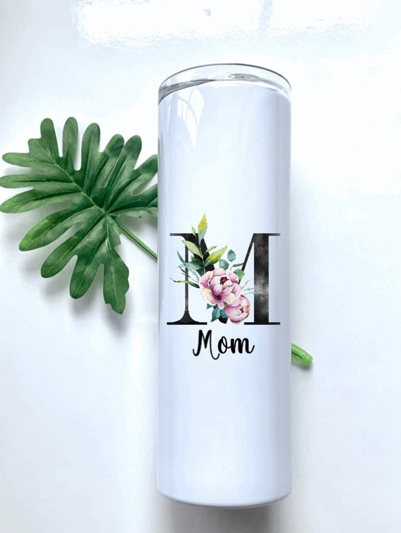 May include: White stainless steel tumbler with a black letter "M" decorated with pink and green floral accents. The word "Mom" is printed below the letter.