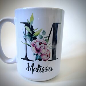 May include: White ceramic mug with a black letter M decorated with watercolor flowers and greenery. The name Melissa is written in black cursive below the letter.