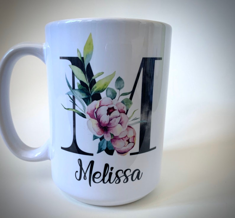 May include: White ceramic mug with a black letter M decorated with watercolor flowers and greenery. The name Melissa is written in black cursive below the letter.