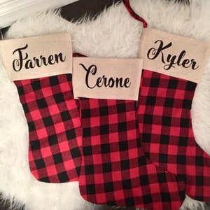 May include: Three Christmas stockings featuring a red and black buffalo plaid pattern. Each stocking has a tan burlap cuff at the top. The names Farren, Cerone, and Kyler are printed in black cursive script on the cuffs of the three stockings.