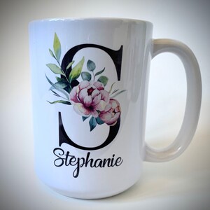 May include: White ceramic mug with a black letter "S" decorated with watercolor pink flowers and green leaves. The name "Stephanie" is written in black cursive below the letter.