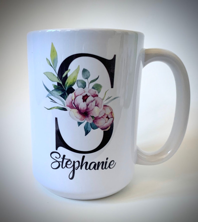 May include: White ceramic mug with a black letter "S" decorated with watercolor pink flowers and green leaves. The name "Stephanie" is written in black cursive below the letter.