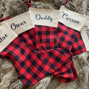 May include: Five Christmas stockings featuring a red and black buffalo plaid pattern with a tan linen cuff. Each cuff is personalized with black script text reading Chloe, Oscar, Daddy, Carson, and Mummy. Each stocking has a red and black plaid loop.