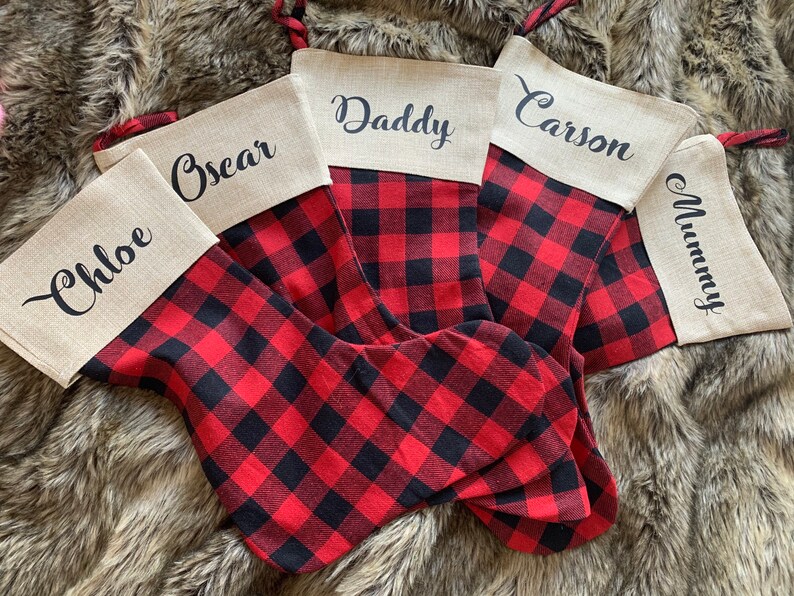 May include: Five Christmas stockings featuring a red and black buffalo plaid pattern with a tan linen cuff. Each cuff is personalized with black script text reading Chloe, Oscar, Daddy, Carson, and Mummy. Each stocking has a red and black plaid loop.