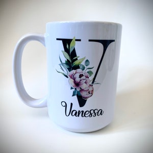 May include: White ceramic mug with a black letter V decorated with watercolor flowers and green leaves. The name Vanessa is written below the letter.