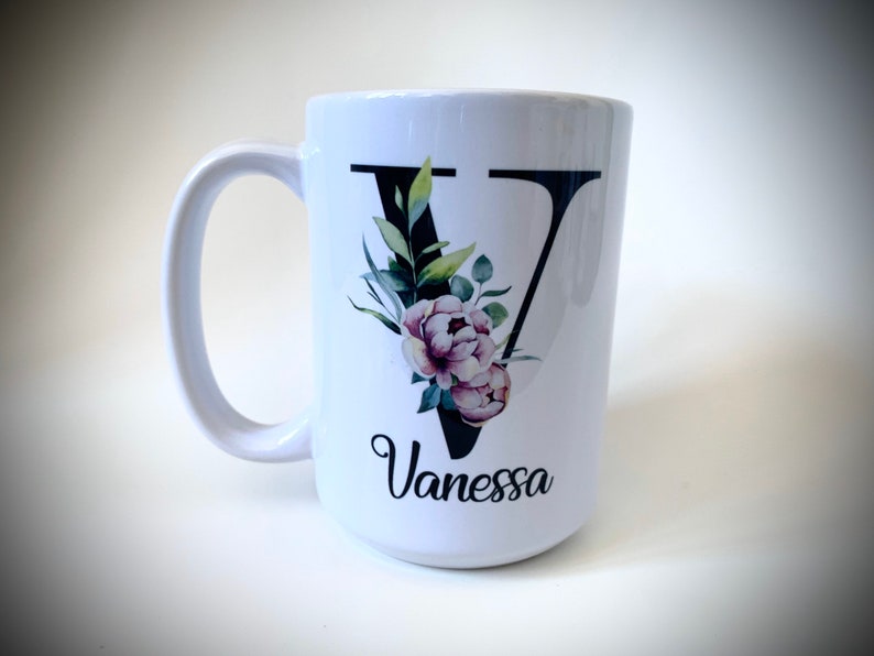 May include: White ceramic mug with a black letter V decorated with watercolor flowers and green leaves. The name Vanessa is written below the letter.