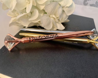 May include: Three diamond-shaped pens with a rose gold, silver, and gold finish. The rose gold pen has the word "Bridemaid" engraved on it.