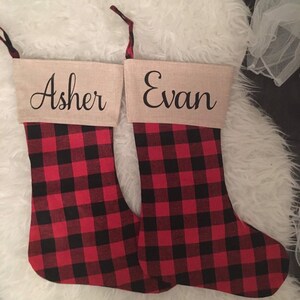 May include: Two Christmas stockings featuring a red and black buffalo plaid pattern on the lower section and a tan cuff at the top. The cuff on the left stocking is personalized with the name Asher, and the right with the name Evan in black script.