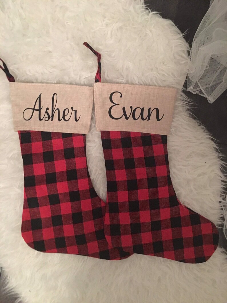 May include: Two Christmas stockings featuring a red and black buffalo plaid pattern on the lower section and a tan cuff at the top. The cuff on the left stocking is personalized with the name Asher, and the right with the name Evan in black script.