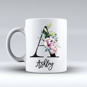 Peut inclure: Une tasse en céramique blanche avec une lettre A noire décorée de fleurs aquarelle et de feuilles vertes. Le nom Ashley est écrit en cursive sous la lettre.