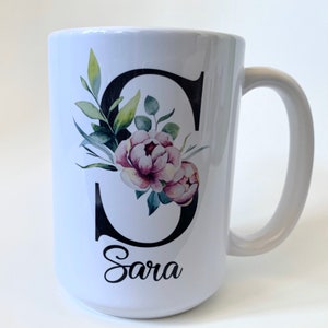May include: White ceramic mug with a black letter "S" decorated with watercolor floral designs in shades of green and pink. The name "Sara" is written in black cursive below the letter.