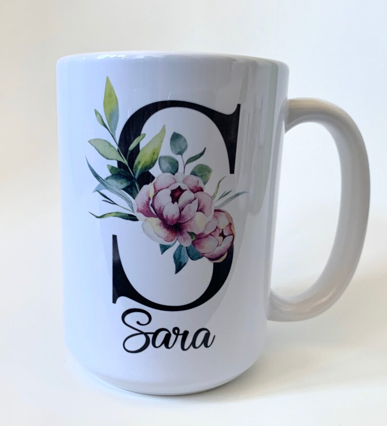 May include: White ceramic mug with a black letter "S" decorated with watercolor floral designs in shades of green and pink. The name "Sara" is written in black cursive below the letter.