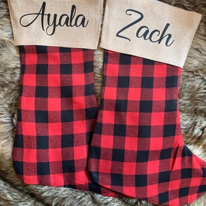 May include: Two Christmas stockings featuring a red and black buffalo plaid pattern on the lower section and a tan burlap cuff at the top. The left stocking is personalized with the name Ayala and the right stocking with the name Zach in black script.