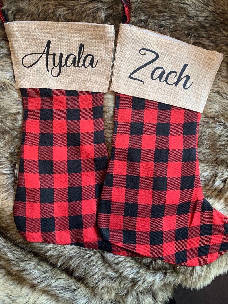 May include: Two Christmas stockings featuring a red and black buffalo plaid pattern on the lower section and a tan burlap cuff at the top. The left stocking is personalized with the name Ayala and the right stocking with the name Zach in black script.
