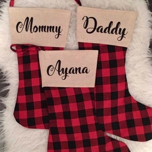 May include: Three Christmas stockings featuring a red and black buffalo plaid pattern. Each stocking has a beige cuff made of a burlap-like fabric. The cuffs are personalized with black script text reading Mommy, Daddy, and Ayana.