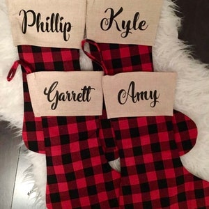 May include: Four Christmas stockings featuring a red and black buffalo plaid pattern. Each stocking has a beige cuff made of a textured fabric. The names Phillip, Kyle, Garrett, and Amy are printed in black cursive script on the cuffs.