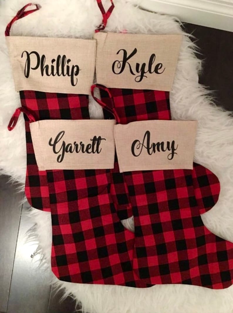 May include: Four Christmas stockings featuring a red and black buffalo plaid pattern. Each stocking has a beige cuff made of a textured fabric. The names Phillip, Kyle, Garrett, and Amy are printed in black cursive script on the cuffs.