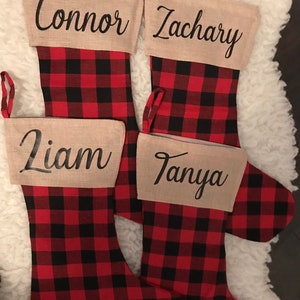 May include: Four Christmas stockings featuring a red and black buffalo plaid pattern. Each stocking has a tan cuff with a name printed in black script: Connor, Zachary, Liam, and Tanya. Each stocking includes a matching plaid hanging loop.