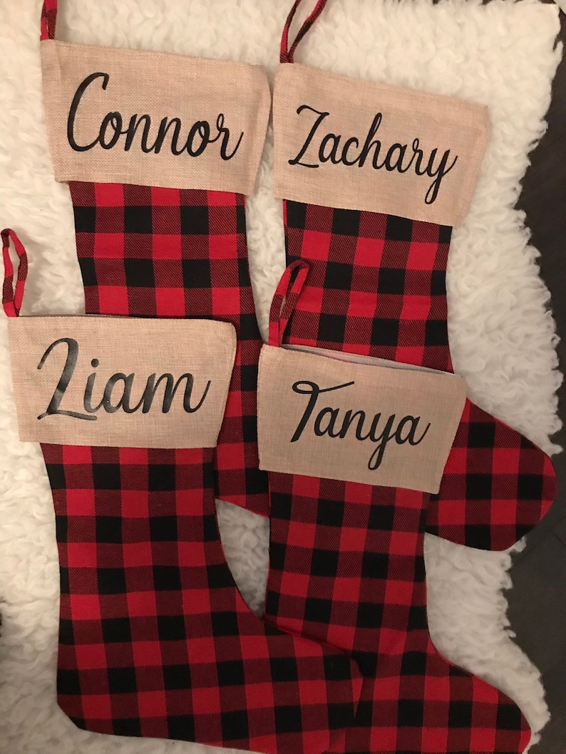 May include: Four Christmas stockings featuring a red and black buffalo plaid pattern. Each stocking has a tan cuff with a name printed in black script: Connor, Zachary, Liam, and Tanya. Each stocking includes a matching plaid hanging loop.