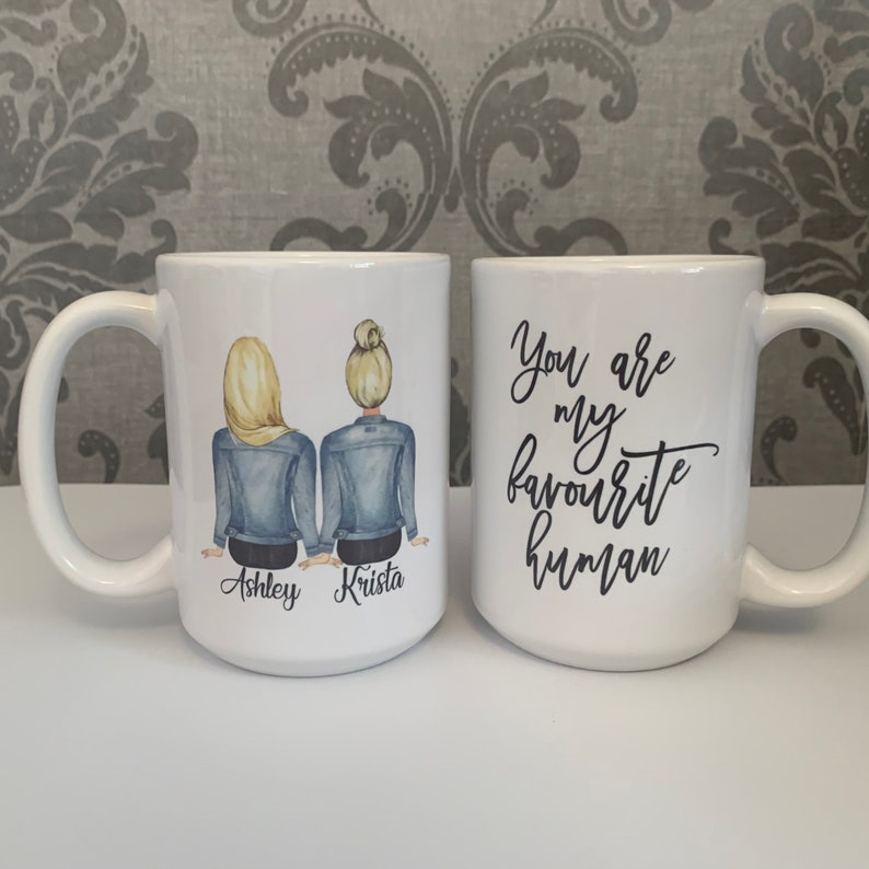May include: Two white ceramic mugs with black handles. One mug features a drawing of two women with blonde hair wearing denim jackets. The text "Ashley Krista" is printed below the women. The other mug has the text "You are my favourite human" printed on it.