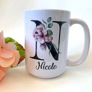 May include: White ceramic mug with a black letter N decorated with pink and green watercolor flowers. The mug has a black handle and the name "Nicole" is written below the letter.