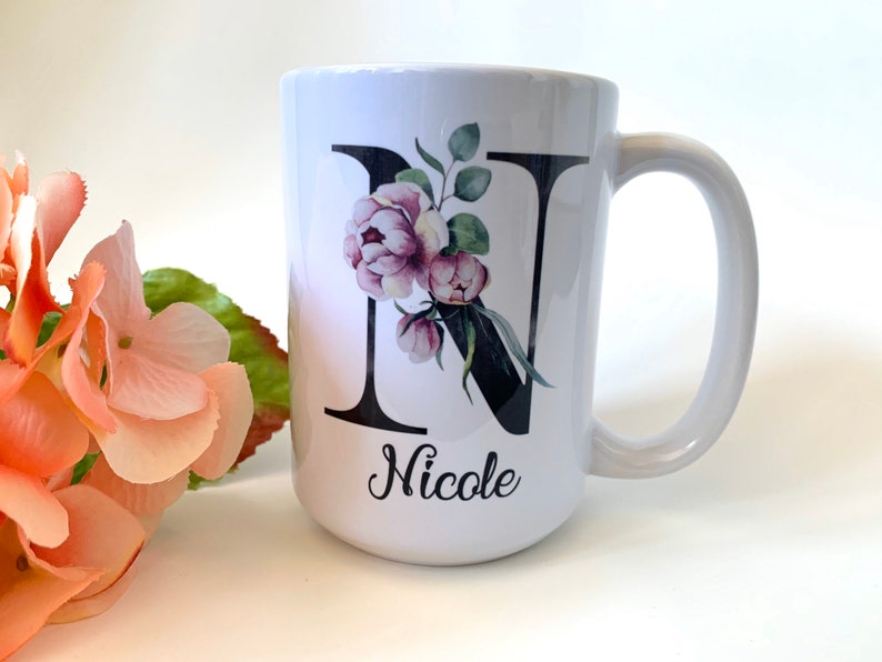 May include: White ceramic mug with a black letter N decorated with pink and green watercolor flowers. The mug has a black handle and the name "Nicole" is written below the letter.