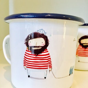 Könnte beinhalten: Eine weiße Emailletasse mit blauem Rand, die eine Cartoon-Illustration eines Mannes mit einem rot-weiß gestreiften Hemd und Bart zeigt. Der Mann hält ein Seil, an dem ein kleiner Fisch befestigt ist.
