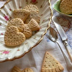 Könnte beinhalten: Ein weißer Keramikteller mit goldfarbenem Rand, gefüllt mit verschiedenen herzförmigen und fischförmigen Shortbread-Keksen. Die Kekse sind mit Puderzucker bestäubt und haben eine hellbraune Farbe. Der Teller liegt auf einer weißen Tischdecke mit Blumenmuster.