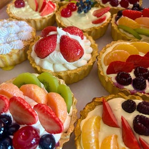 Puede incluir: Una colección de tartaletas de frutas con bases doradas y rellenos cremosos. Cada tartaleta está cubierta con una variedad de frutas frescas, incluyendo fresas, frambuesas, arándanos, kiwi y mandarinas. Las tartaletas están dispuestas sobre una superficie clara.