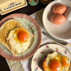 May include: A breakfast scene with two plates of toast topped with fried eggs. A butter dish with the word "LURPAK" is visible, along with decorative figurines and a bowl of eggs. The plates have floral designs. The scene is set on a table.
