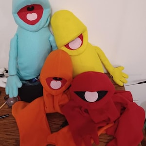 May include: Four plush hand puppets with open mouths and pink tongues. The puppets are blue, yellow, orange, and red. The puppets are sitting on a wooden surface.
