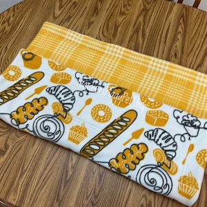 May include: A white and yellow pet bed with a bakery-themed design. The bed features illustrations of bread, croissants, donuts, and cupcakes in yellow and black. The top layer has a yellow plaid pattern.
